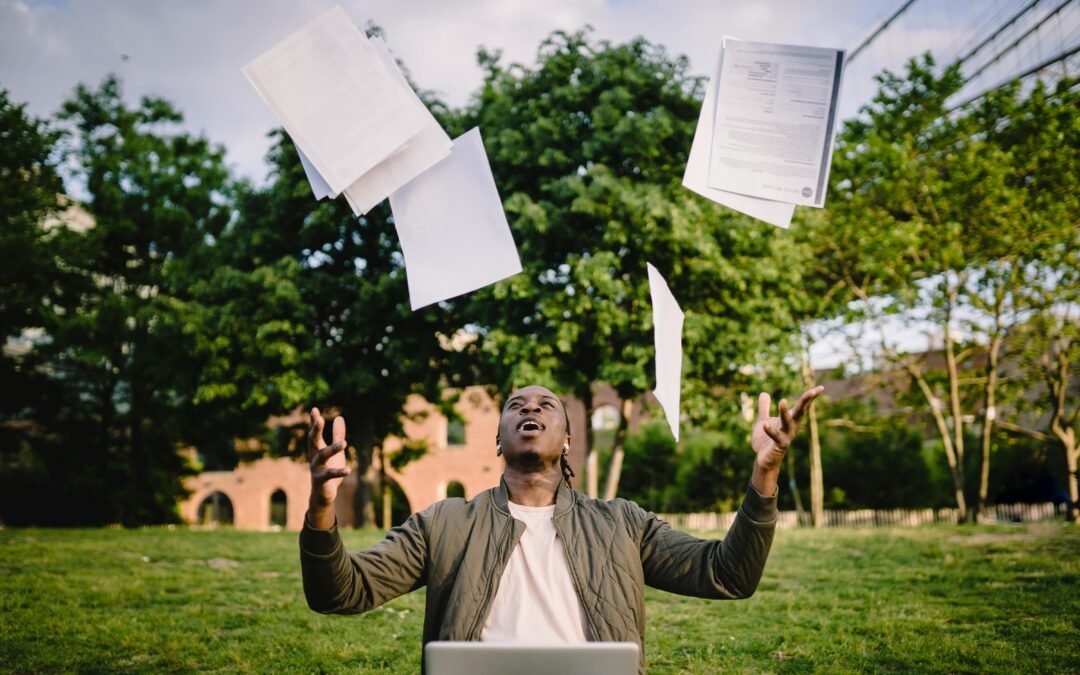 Overjoyed African American graduate tossing copies of resumes in air after learning news about successfully getting job while sitting in green park with laptop